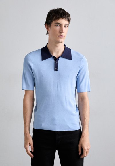 Polo shirt bleu clair à manches courtes avec un col et un plastron de boutonnage bleu marine foncé, présentant un motif en tricot texturé. Porté avec un pantalon noir.