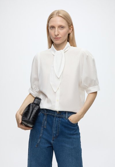 Femme aux cheveux blonds raides portant un chemisier blanc à manches bouffantes avec un col noué et un jean bleu, tenant un sac à main noir texturé.