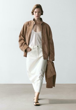 Femme marchant en jupe blanche, débardeur blanc, veste en daim marron clair tenant un manteau assorti et portant des sandales noires.