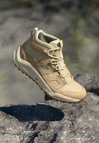 Beige túracipő közepes szárú kialakítással. Texturált felsőrésszel, megerősített orral és gumi talppal rendelkezik, amely tapadást biztosító mintázattal van ellátva.