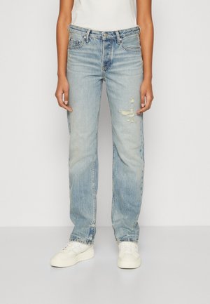 Personne portant un jean droit déchiré bleu clair et des baskets blanches, debout devant un fond uni clair.