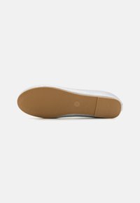 Semelle de sneaker blanche avec des rainures texturées et une plateforme en caoutchouc marron clair. Présente une empreinte de logo rond près du talon.