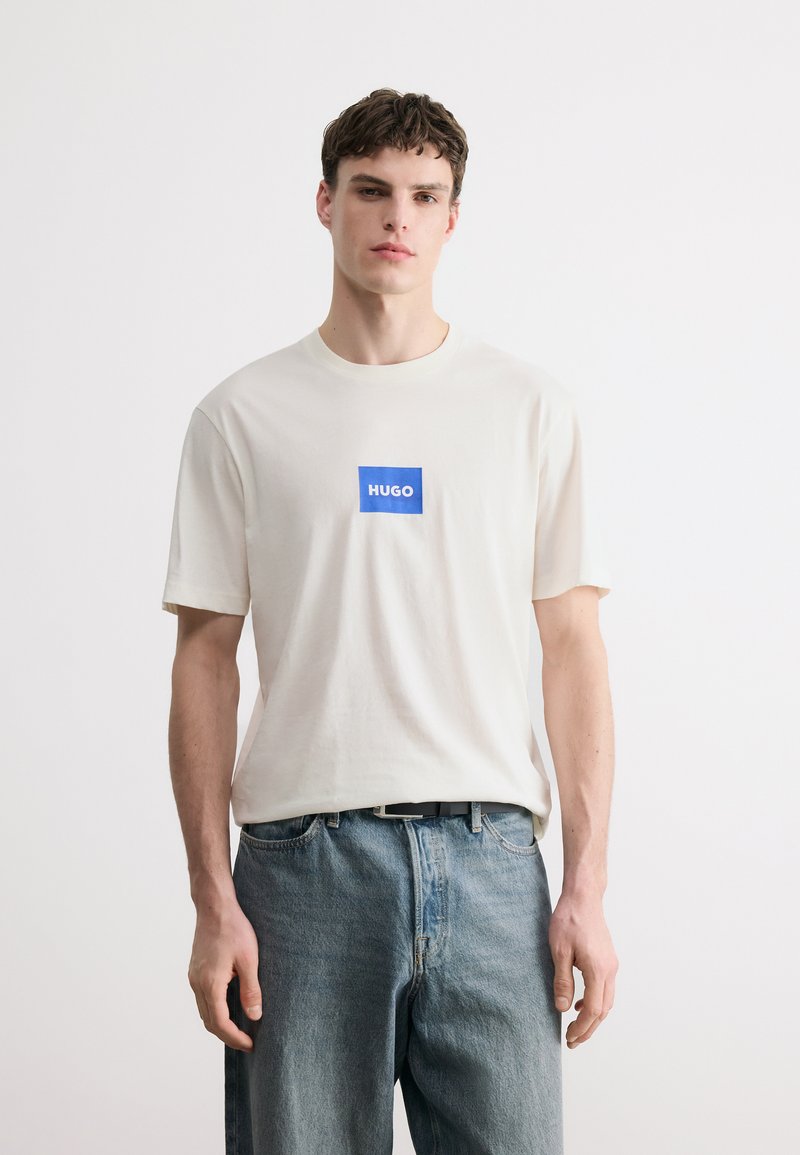 Νεαρός άνδρας που φοράει λευκό μπλουζάκι με μπλε λογότυπο «HUGO» και ανοιχτόχρωμο μπλε τζιν, στέκεται μπροστά σε απλό λευκό φόντο.