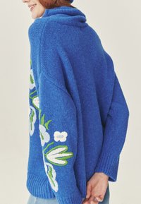 Pulover tricotat albastru cu glugă, având broderie florală albă și verde pe mânecă. Material texturat și croială relaxată.