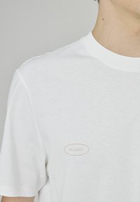 T-shirt en coton blanc avec un col rond simple, des manches courtes, et une étiquette « RELAXED » rose clair sur la poitrine gauche.