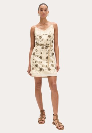 Kvinde iført en beige ærmeløs blomsterkjole med et tyndt bælte og brune remsandaler, stående på en lys baggrund.