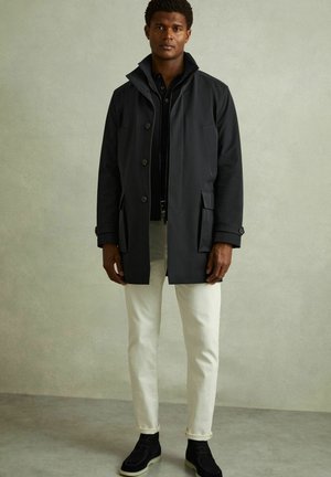 Cappotto impermeabile grigio scuro con colletto alto, chiusura a bottoni e tasche frontali, abbinato a jeans bianchi e stivali alla caviglia scuri.