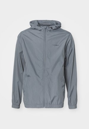 Giacca a vento leggera grigia con cappuccio, polsini elasticizzati e tasche laterali. Realizzata in materiale liscio e resistente all'acqua.