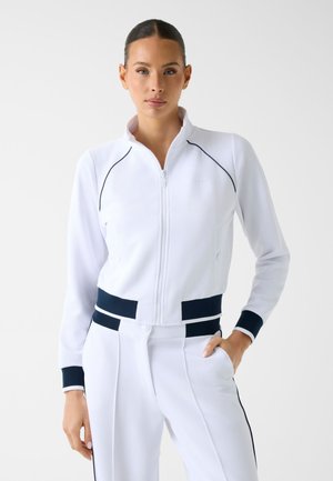 Frau mit dunklen Haaren in einer weißen Jacke mit Reißverschluss und passenden weißen Hosen mit schwarzem Besatz, stehend vor einem einfachen hellen Hintergrund.