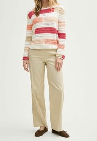 Femme portant un pull en tricot rayé multicolore, un pantalon beige à jambes larges et des ballerines marron, debout devant un fond blanc.