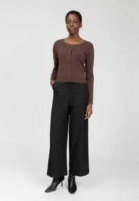 Braune taillierte Strickjacke mit Knöpfen, kombiniert mit hoch taillierten schwarzen weit geschnittenen Hosen und spitzen schwarzen Absätzen, steht vor einem neutralen Hintergrund.