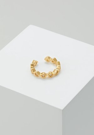 Bague ouverte en or avec des motifs en forme de diamant, chacun serti de petites pierres claires, présentée sur une plateforme blanche inclinée.