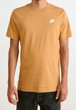 Man draagt een mosterdgele Nike T-shirt met korte mouwen en zwarte broek, staand tegen een effen lichte achtergrond.