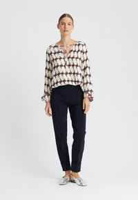 Lange mouwen blouse in crème met bordeaux geometrisch patroon, v-hals met trekkoord; marineblauwe slim-fit broek; zilveren platte schoenen.