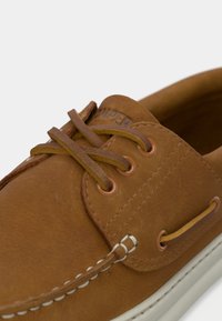 Καφέ δερμάτινο παπούτσι τύπου boat με κοντράστ ραφές, χαρακτηριστικά μπρούτζινα τρύπες για τα κορδόνια, κλείσιμο με κορδόνια και λευκή ελαστική σόλα.
