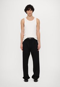 White sleeveless tank top and loose black trousers. Trousers have an elastic waistband with a logo. Model stands against a plain background.