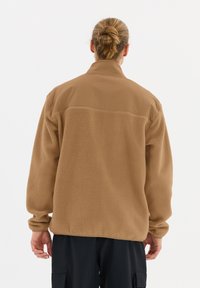 Veste en polaire marron avec un corps texturé, un panneau d'épaule lisse, un col haut et des poignets élastiques. Portée sur un pantalon noir, vue de dos.