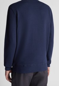 Osoba oblečená v jednobarevné tmavě modré mikině s dlouhým rukávem a tmavých kalhotách, zobrazená zezadu na neutrálním pozadí.