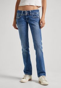 Helle blaue Denim-Jeans mit schlankem Schnitt, mittelhoher Taille, zwei Gesäßtaschen und doppeltem Knopfverschluss. Getragen mit weißen Sneakers.