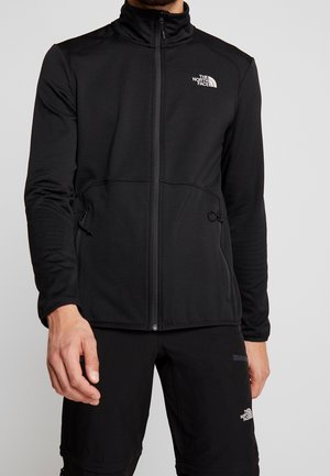 Mann trägt schwarze Fleecejacke mit Reißverschluss und schwarze Outdoorhose, beide mit The North Face-Logo auf linker Brust- und Oberschenkelseite.