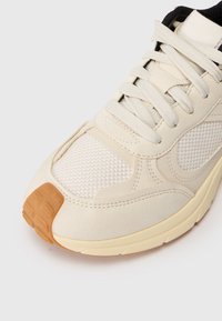 Zapatilla deportiva beige con parte superior de malla y ante, puntera redondeada, cordones planos y suela de goma. Presenta acentos en marrón claro y detalles texturizados.