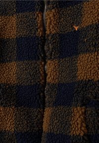 Chaqueta de forro polar con cremallera que presenta un patrón a cuadros en azul oscuro y marrón, con una superficie suave y texturizada y un detalle del logotipo en naranja que contrasta.