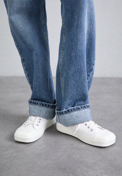 Vita canvas sneakers med en tjock gummisula, utrustade med metalöglor och platta snörningar, ihop med blå denimjeans som är uppvikta vid bensluten.