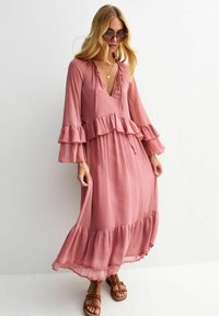 Lång, rosa klänning med volangärmar och fåll. Lättviktsmaterial, V-ringning och dragsko i midjan. Stylistiskt kombinerad med bruna sandaler med remmar.