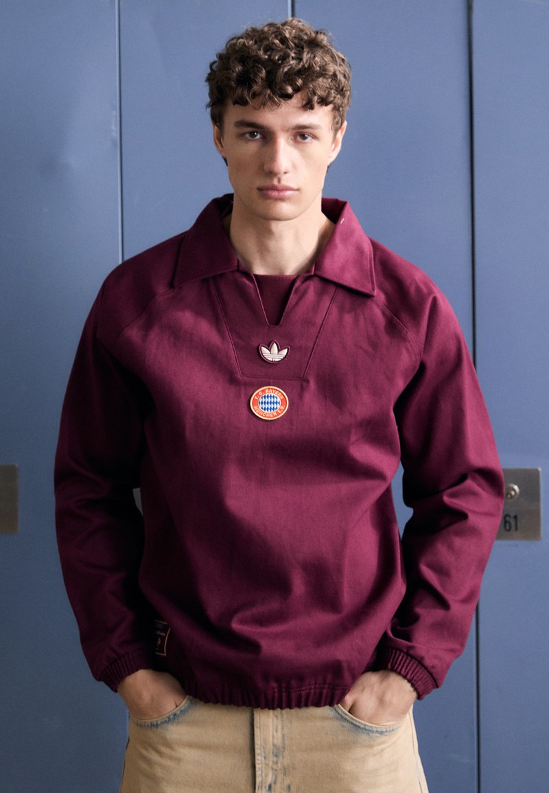 Pull maroon avec un large col, comportant un petit logo Adidas et un écusson circulaire sur la poitrine. Tissu texturé, poignets côtelés.