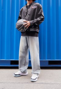 Black satin bomber jacket with white stripes, graphic tee, light-wash wide jeans, and white sneakers holding a black basketball against a blue background.