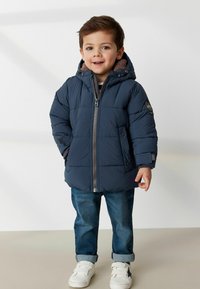 Marineblaue Steppjacke mit Reißverschluss, Kapuze, strukturiertem Futter und Logo-Patch. Kombiniert mit hochgekrempelten Jeans und weißen Sneakers.