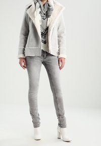Veste en fausse fourrure grise avec un col en shearling, écharpe à motifs noir et blanc, jean skinny gris et bottines blanches à talons.