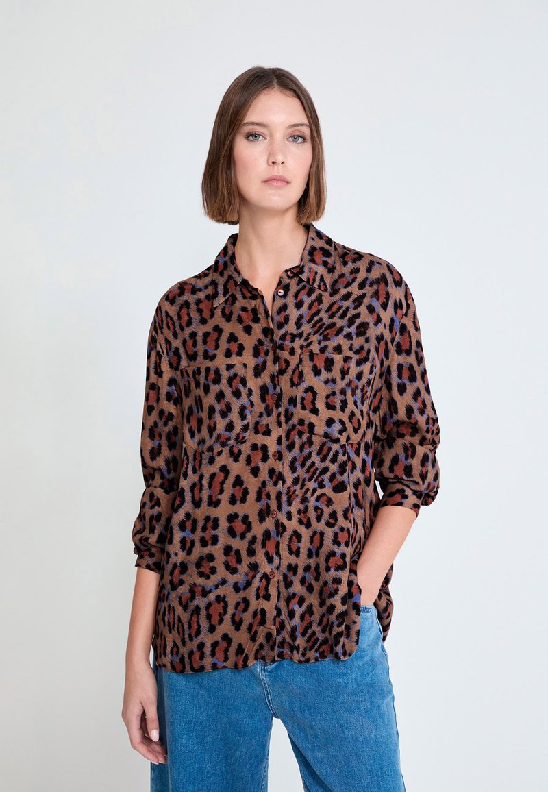 Blouse imprimé léopard en brun, avec une texture douce, une fermeture à boutons sur le devant, des manches longues, et une poche poitrine. Portée avec un jean bleu.