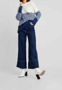 Chunky stickad tröja i kräm och blå, med en texturerad design; ihop med högmidjade, vida mörk denimjeans och vita klackstövlar.