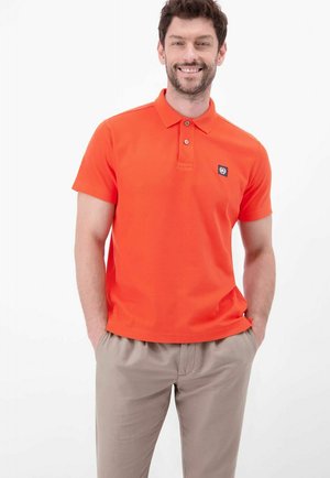 Mężczyzna stoi z rękami w kieszeniach, ubrany w pomarańczową, krótkorękawową koszulkę polo i beżowe spodnie, uśmiechając się na białym tle.