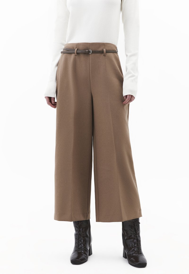 Person trägt hochtaillierte, weit geschnittene braune Hose mit dünnem dunklem Gürtel, weißes langärmeliges Oberteil und schwarze Schnürstiefel mit Absatz.