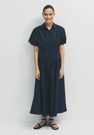 Lächelnde Frau mit dunklen Haaren in einem marineblauen Midikleid mit kurzen Ärmeln, die vor einem schlichten hellen Hintergrund steht und schwarze Sandalen trägt.