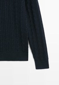 Rukáv a spodní polovina námořnicky modrého svetr s kabelovým vzorem, s žebrovaným lemem a manžetou na bílém pozadí.