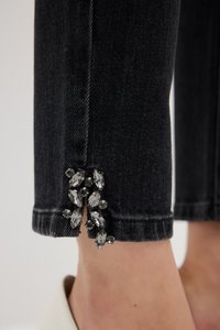 Černé džíny z denimového materiálu mají roztrhaný lem ozdobený tmavými rhinestones, což vytváří texturovaný kontrast v oblasti kotníku.