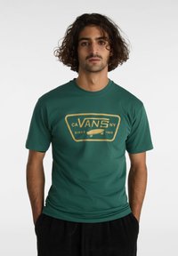 Camiseta verde de manga corta con un diseño gráfico dorado que incluye el texto "CA VANS NY" y una patineta, hecha de material de algodón suave.