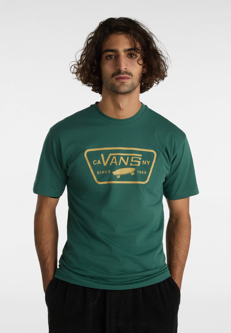 Camiseta verde de manga corta con un diseño gráfico dorado que incluye el texto "CA VANS NY" y una patineta, hecha de material de algodón suave.