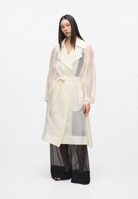 Trench-coat beige clair semi-transparent avec ceinture nouée, manches longues, et accents translucides, porté avec un pantalon large noir transparent.