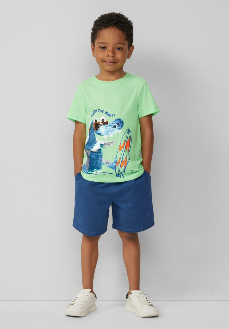 Lichtgroene katoenen t-shirt met een dinosaurusafbeelding en de tekst "Catch the Wave!", gecombineerd met blauwe shorts en witte sneakers.