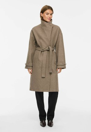 Femme portant un manteau en laine marron ceinturé avec un col montant, un pantalon noir et des bottines foncées, debout devant un fond clair unicolore.