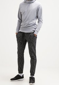 Mann in einem hellgrauen langärmeligen Hoodie, dunkelgrauen, schmal zulaufenden Jogginghosen mit Kordelzug, weißen Socken und schwarzen Sportschuhen, der mit den Händen in den Taschen dasteht.