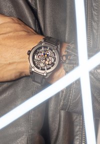 Juodas analoginis laikrodis su matomu mechaniniu mechanizmu, tekstūruota juoda gumos apyrankė ir sidabriniai akcentai. Nešiojamas ant rankos, apsaugotos odine striukė.
