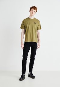Camiseta de algodón verde olivo de manga corta, que presenta un pequeño logo negro de "LEVI'S". Combinada con unos jeans negros ajustados y zapatos negros.