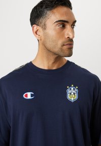 Chemise en coton bleu marine à manches longues avec un logo sur la poitrine, un badge avec des étoiles et un emblème Champion sur le côté gauche.
