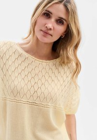 Żółty sweter z dzianiny z wzorem w diamenty, krótki rękaw i luźny krój. Teksturowane ciało z ażurowym stylem na górze.
