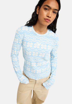 Femme portant une chemise bleu clair à manches longues avec des motifs floraux blancs et un pantalon beige, debout, mains dans les poches.